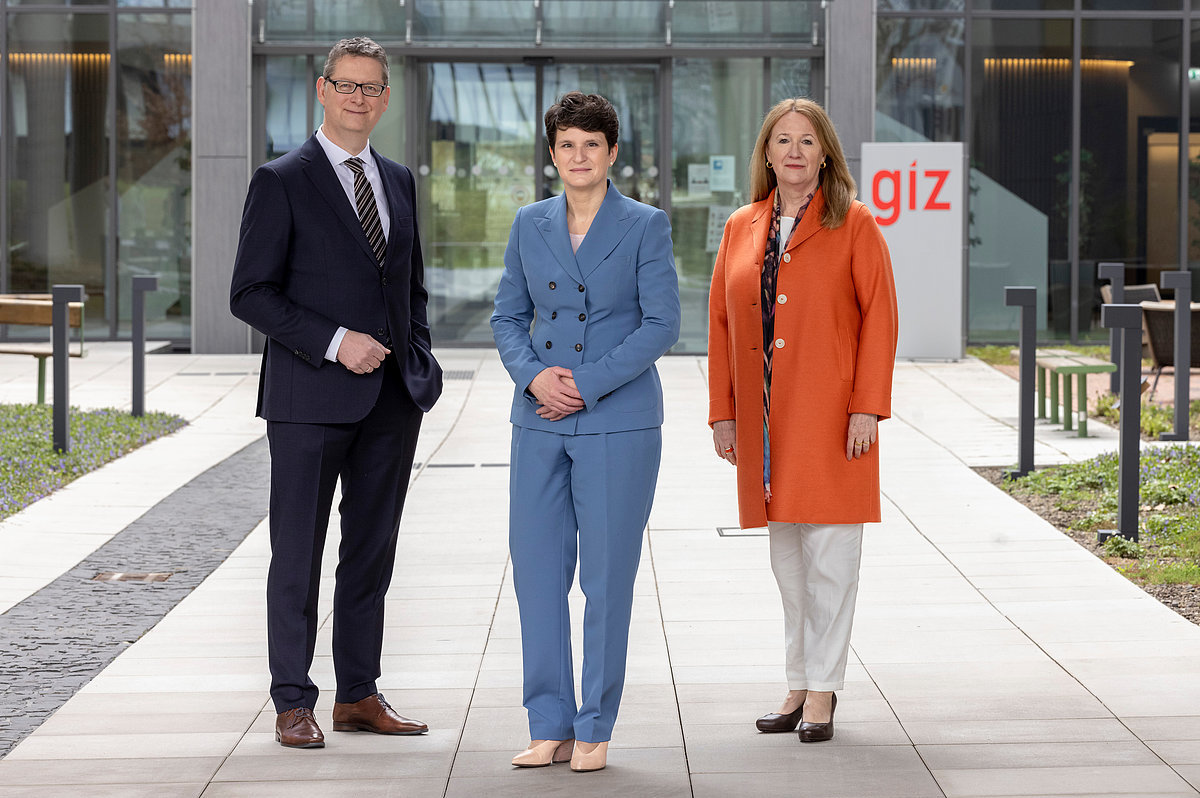Photo: Three people stand slightly apart on a path leading to one of the GIZ buildings.