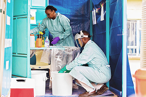 Photo: Two people in a room with walls made of tarpaulins. One person is wearing a transparent plastic visor. Both are wearing gloves and using different disinfectants.