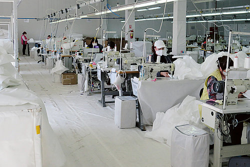 Photo: People sit at industrial sewing machines in a big hall. They are sewing white fabric with white thread.
