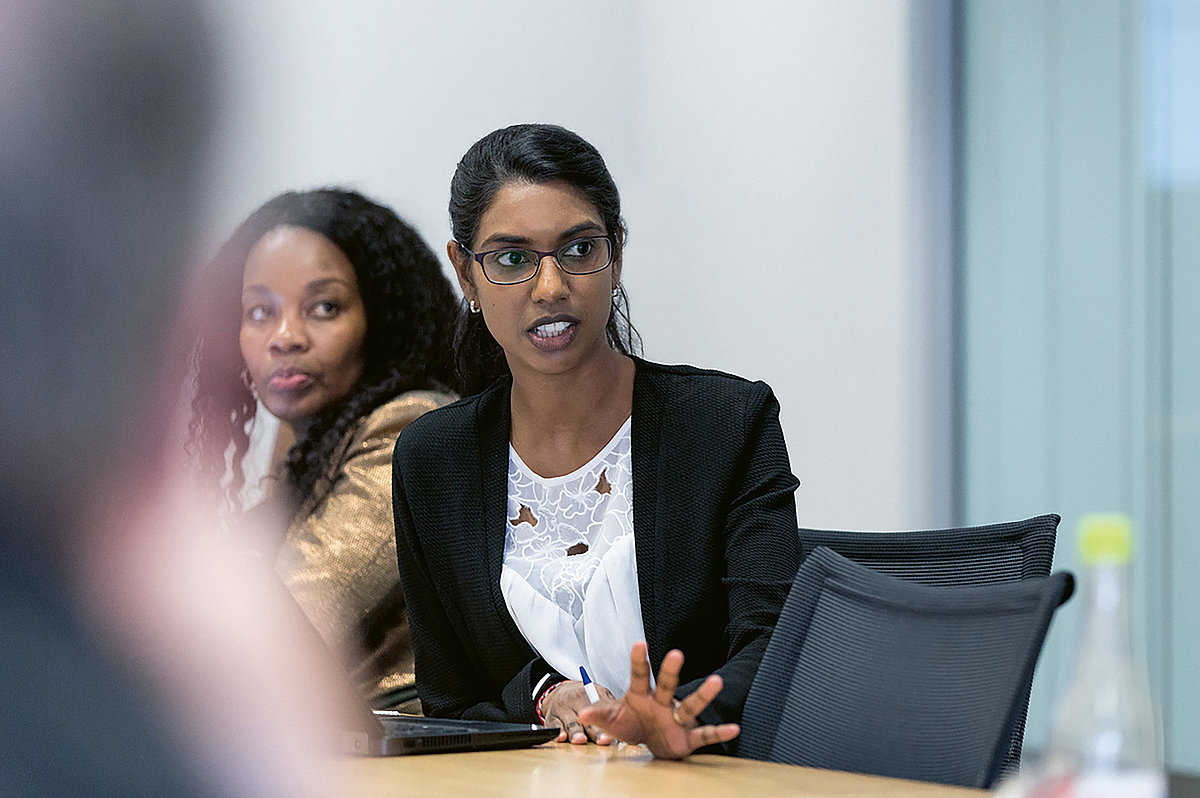 Photo: Two women sit next to each other at a table. One has an open laptop in front of her while she speaks.
