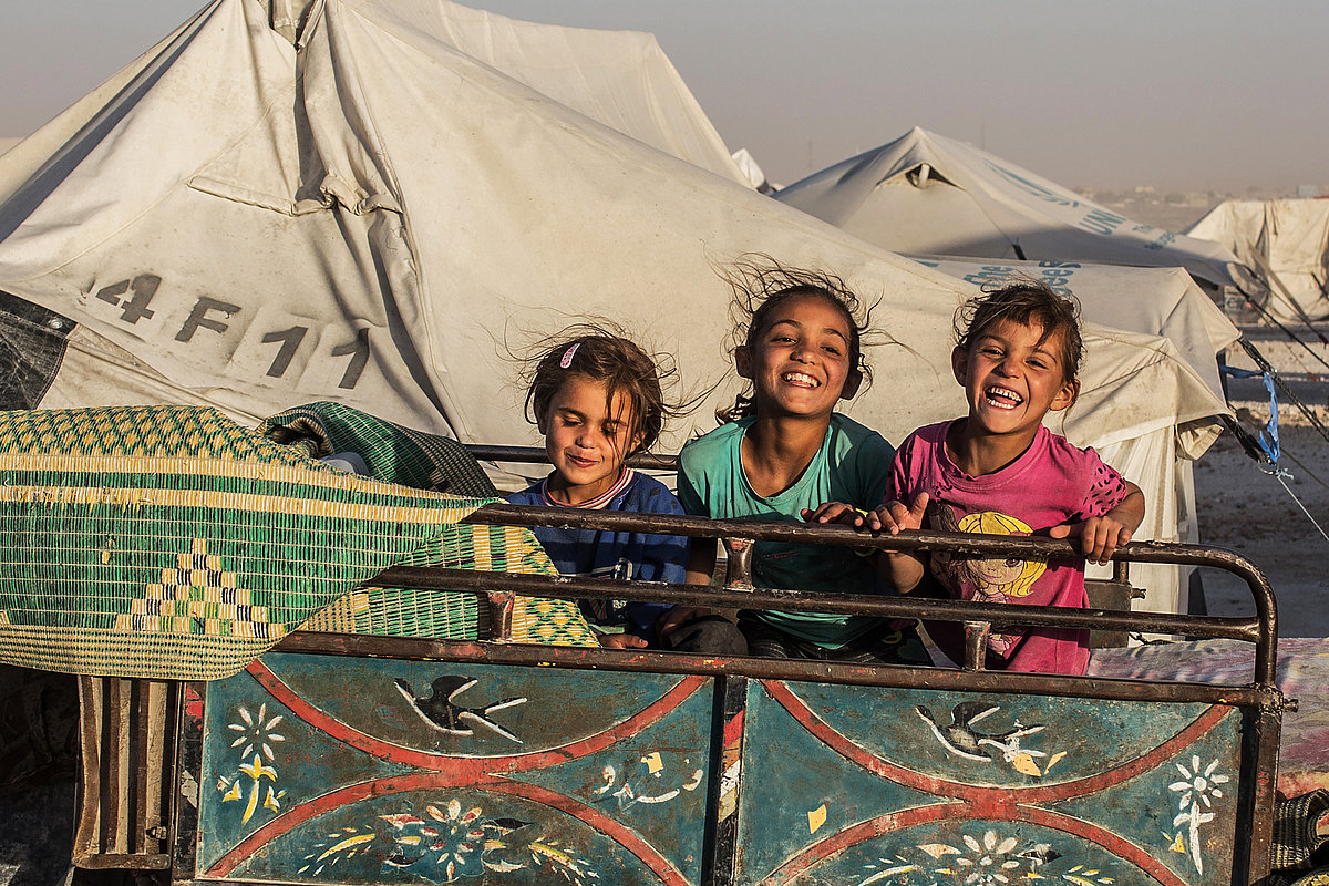 Photo: Three laughing children sit on the back of a lorry behind metal railings. There are large tents in the background.