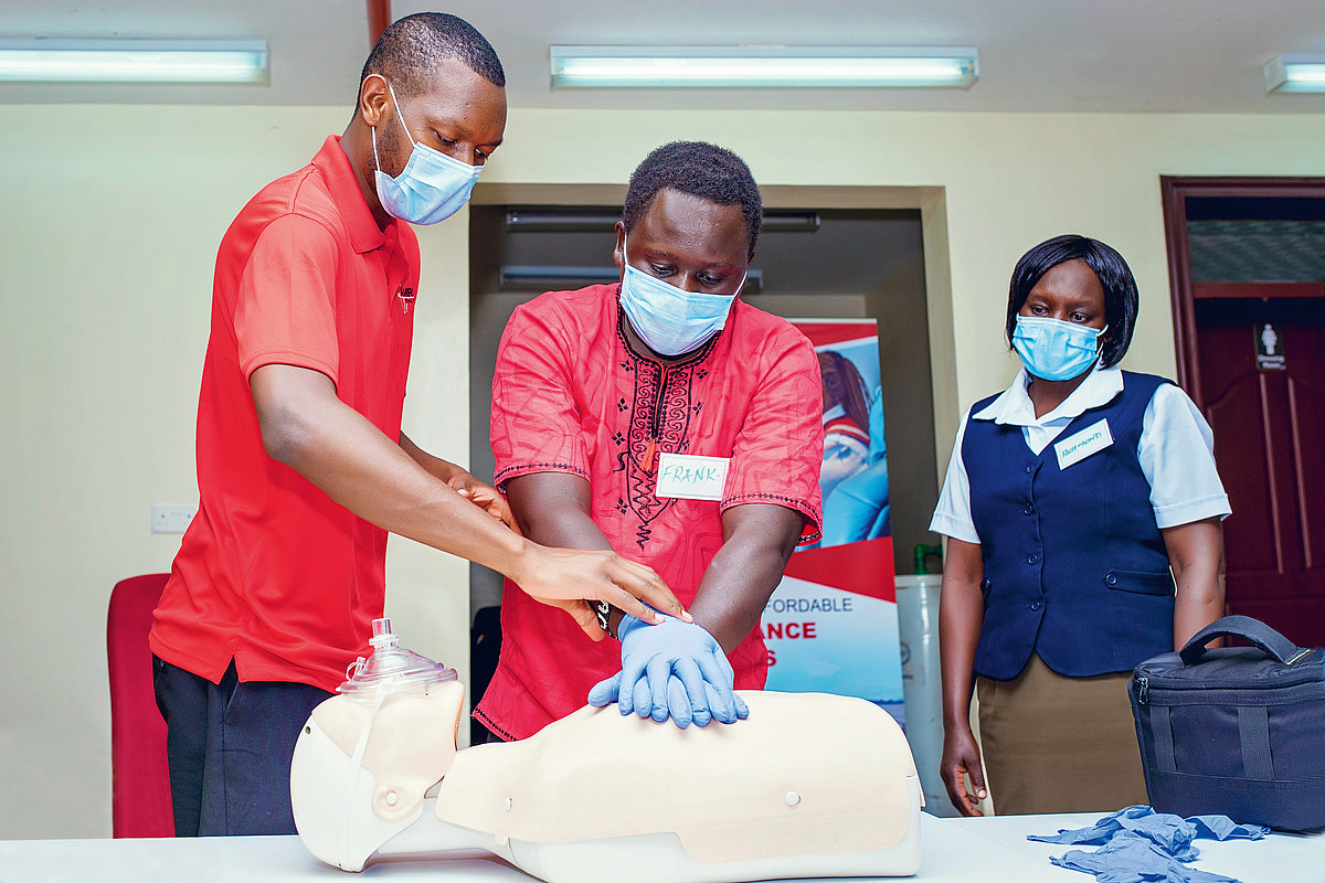 Photo: Three people in a room, all wearing surgical masks. One person with surgical gloves practises heart massage on a CPR doll. Another person explains the technique, while the third person watches in the background.