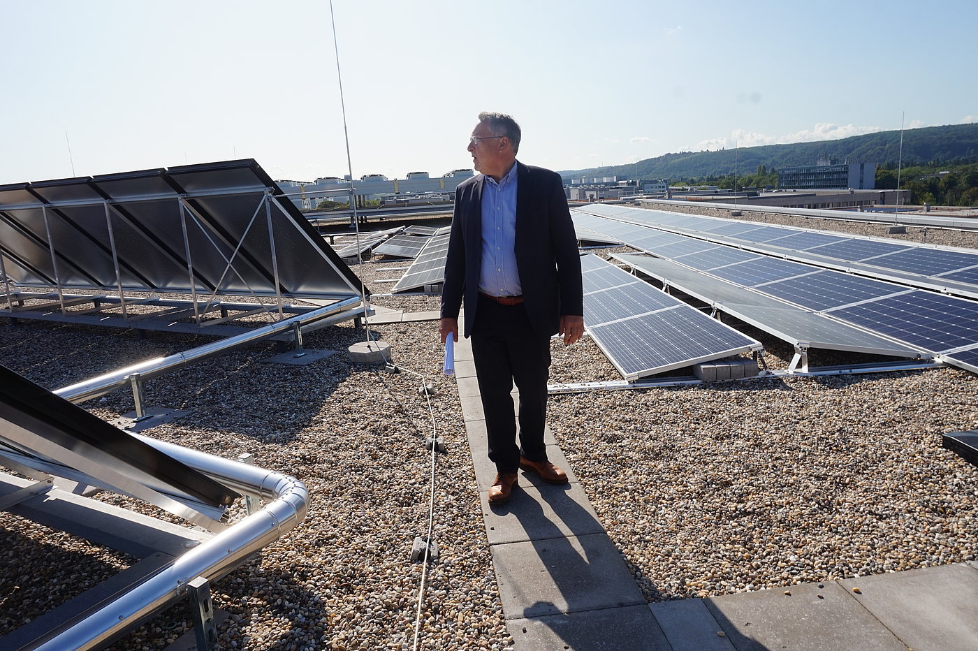Photo: A man stands on paving stones that form paths across gravel. Beside the paths are a large number of photovoltaic panels facing in different directions, linked by pipes.