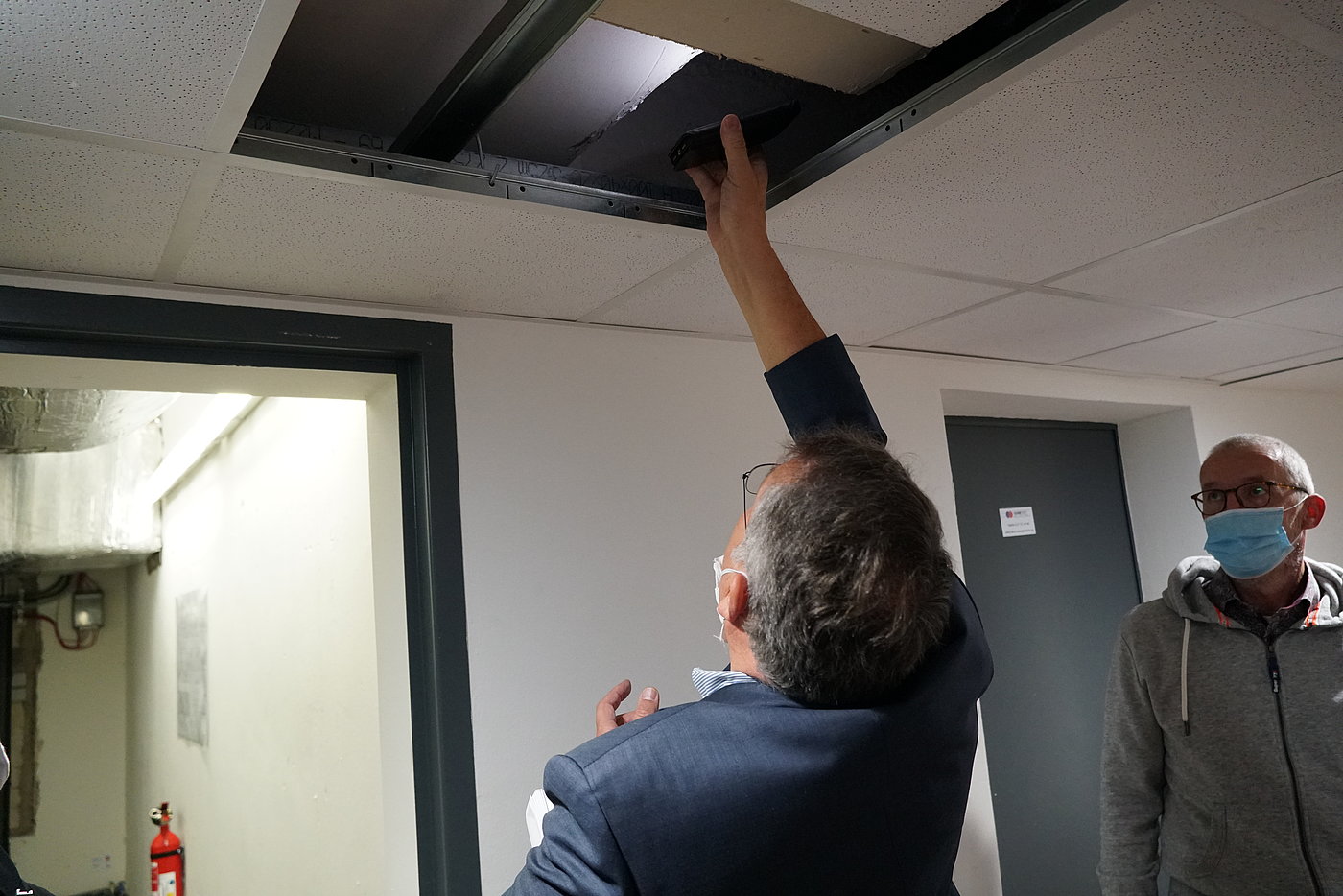 Photo: A person, wearing a surgical mask, reaches into an open cable duct in a suspended ceiling. Another person watches, also wearing a surgical mask.