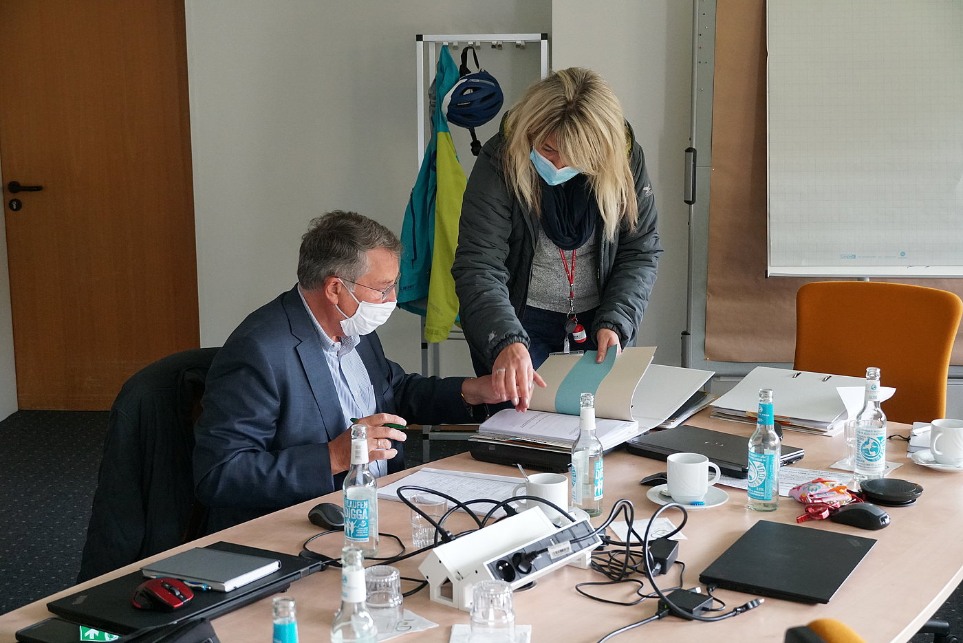 Photo: A man wearing a surgical mask sits at a desk. Other desks beside him form one large area, with electrical connections and devices. A woman, also wearing a surgical mask, stands beside the man, and points to a page in a file. A flipchart can be seen in the background.
