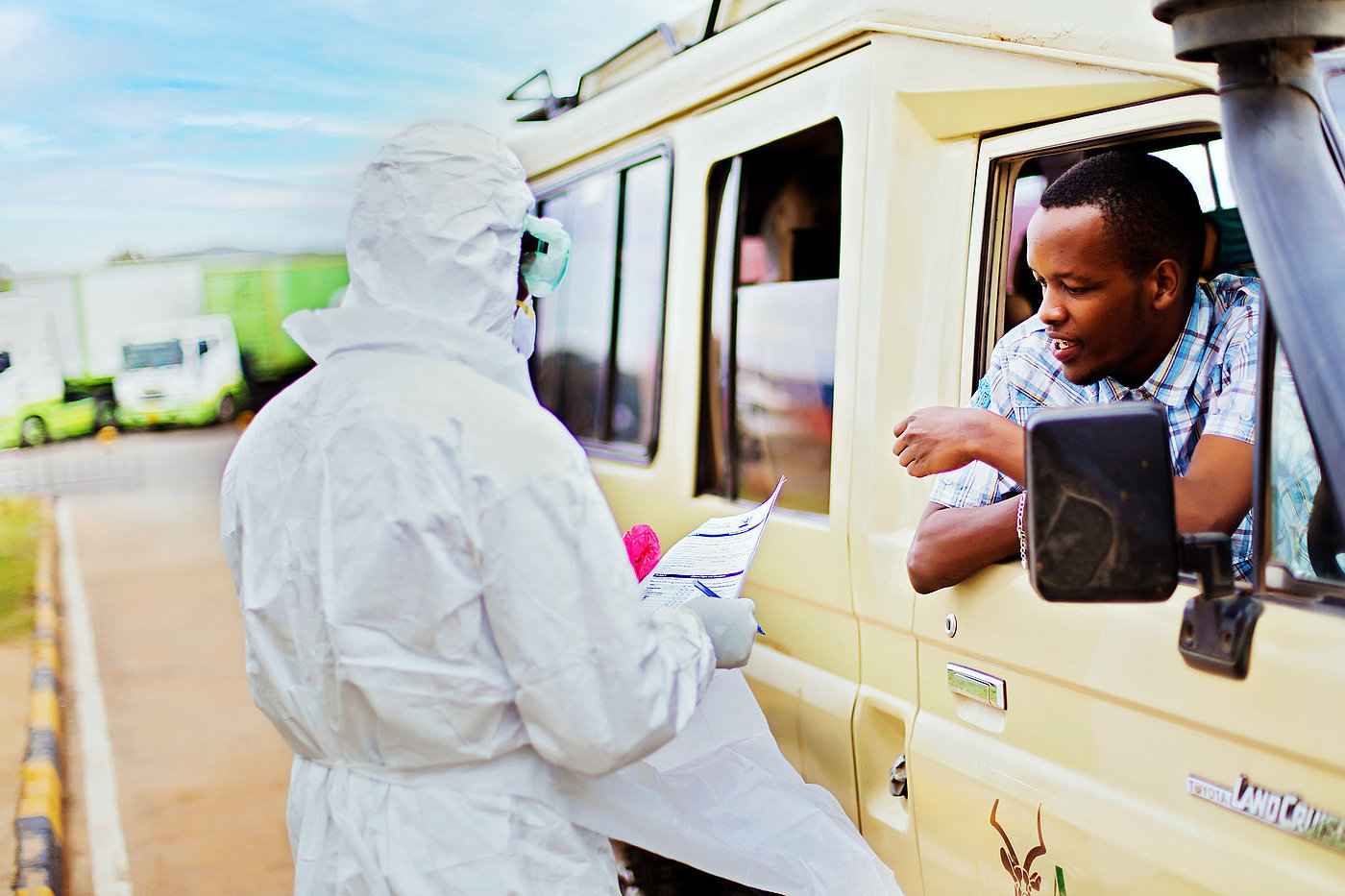 Photo: A person wearing a protective suit holds a document and stands in front of a vehicle. She is speaking to a young man leaning out of the vehicle’s window.
