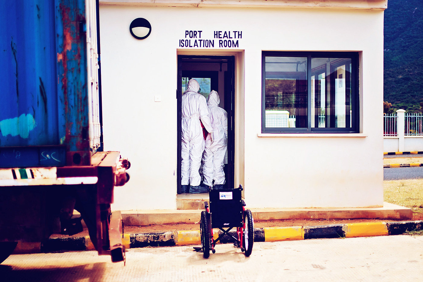 Photo: Two people in protective suits stand in the open entrance to a building. There is a wheelchair in front of the building.