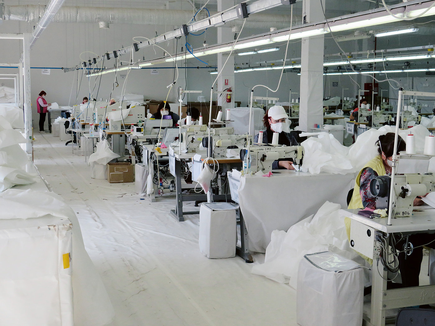 Photo: People sit at industrial sewing machines in a big hall. They are sewing white fabric with white thread.