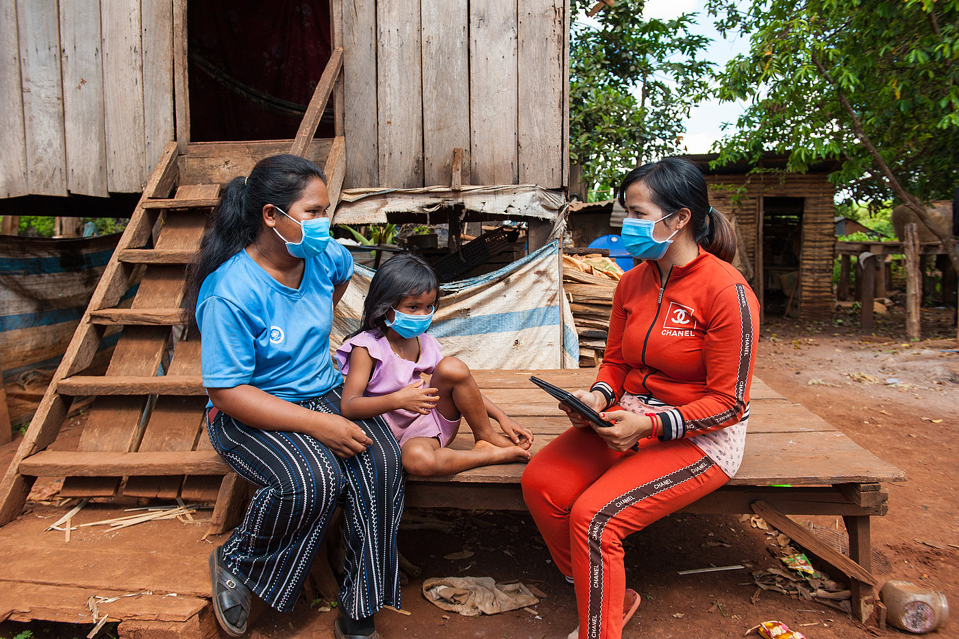 Photo: Two women and a child sit on a wooden platform in front of a wooden house. All of them are wearing surgical masks. One woman is holding a tablet PC with both hands.