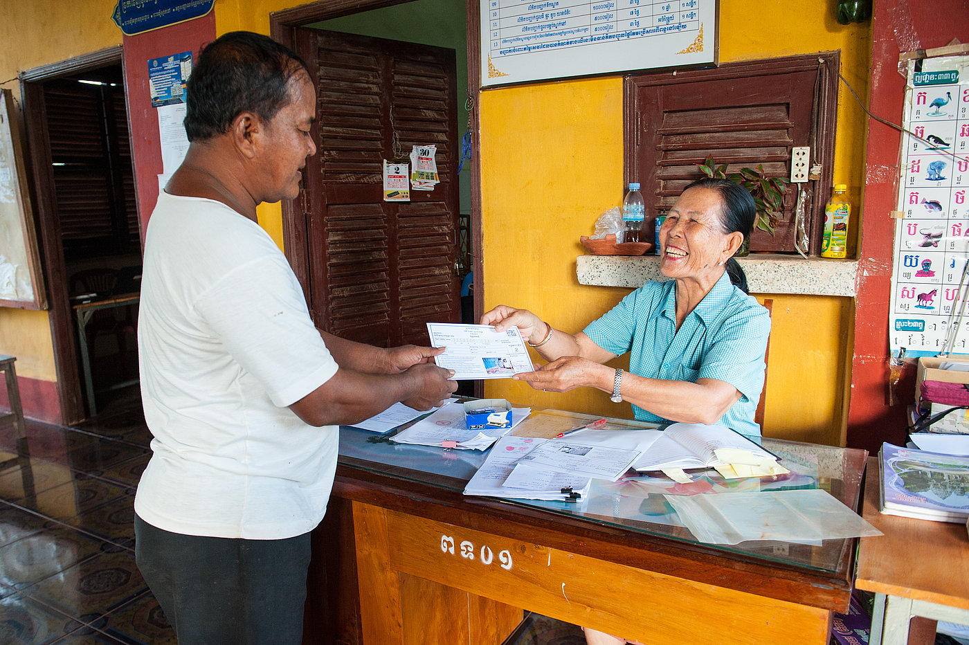 Photo: A man stands in front of a table at which a woman is sitting. The woman smiles and hands him a document.