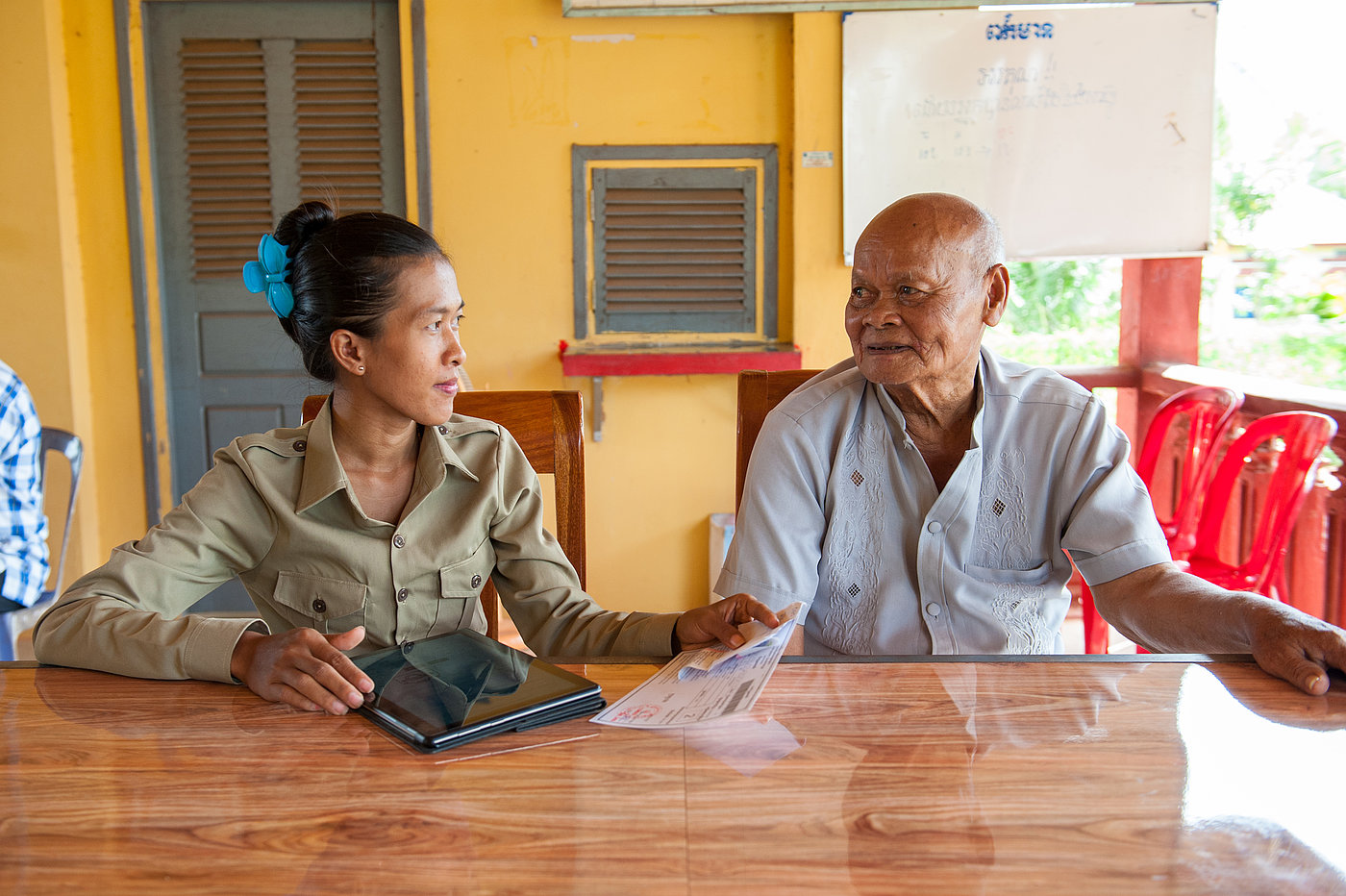 Photo: Two people sit side by side at a table and look at one another.  A document and a tablet PC lie on the table.