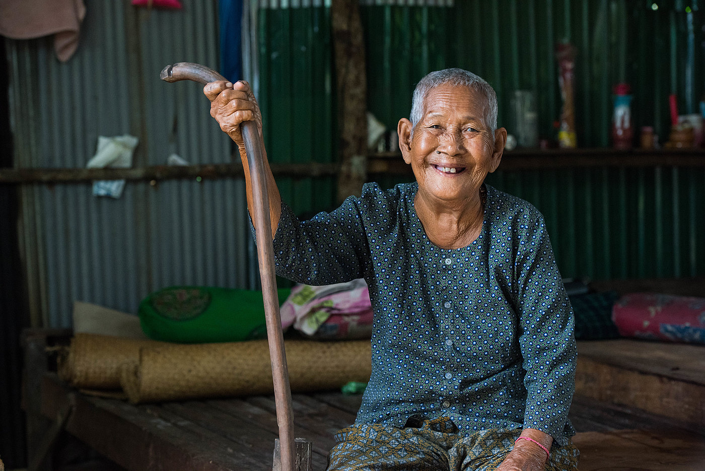 Photo: An elderly woman sits and smiles at the camera. In her right hand she is holding a long stick.