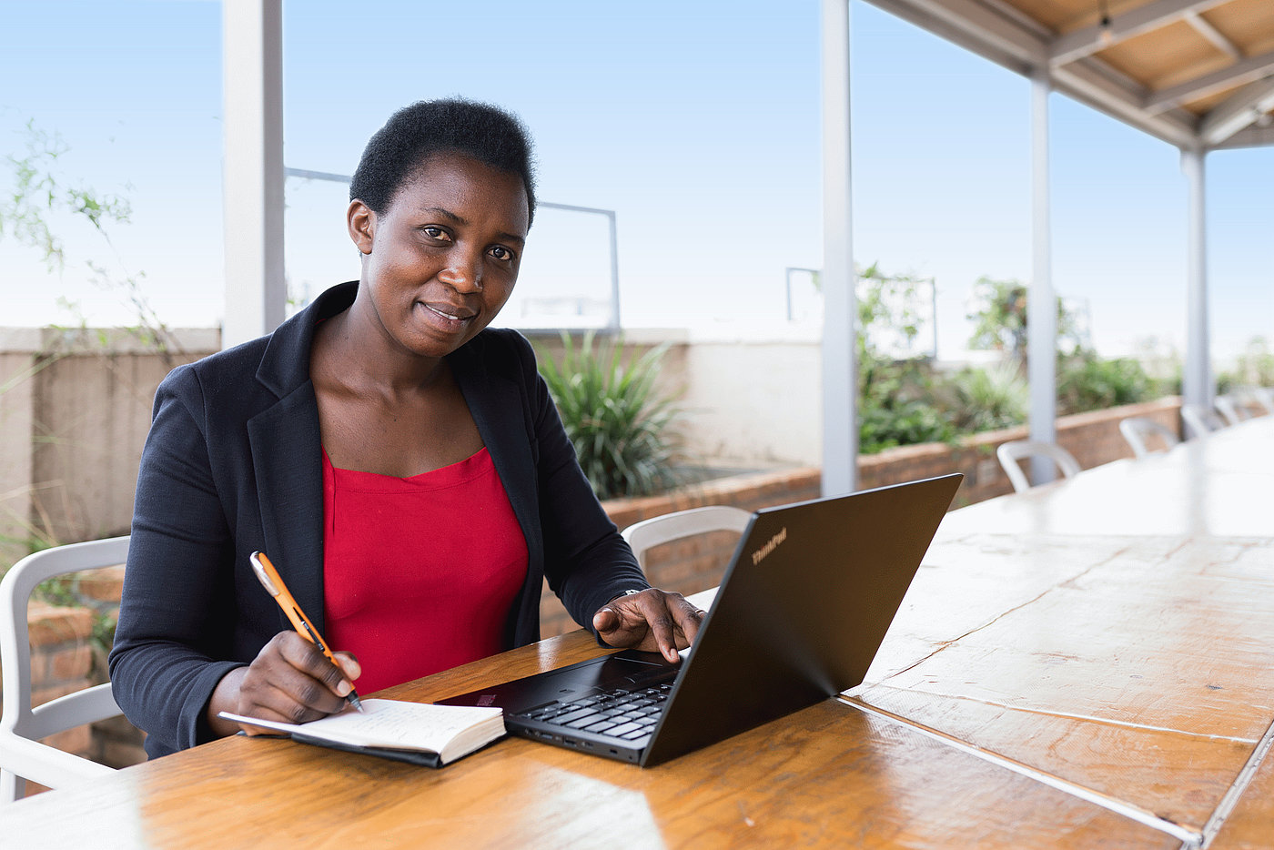 Photo: A woman sits at a desk outside. She has a laptop open in front of her. She is jotting something down in a notebook, which lies beside the laptop on the table.