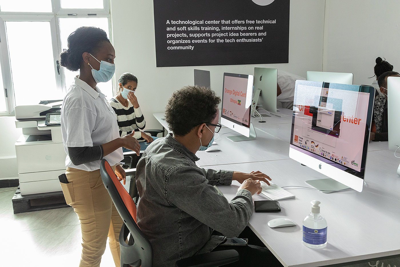 Photo: Three people are in a room with large computer monitors. All of them are wearing surgical masks. Two are sitting at computer workstations, while another person looks over the shoulder of one of them.