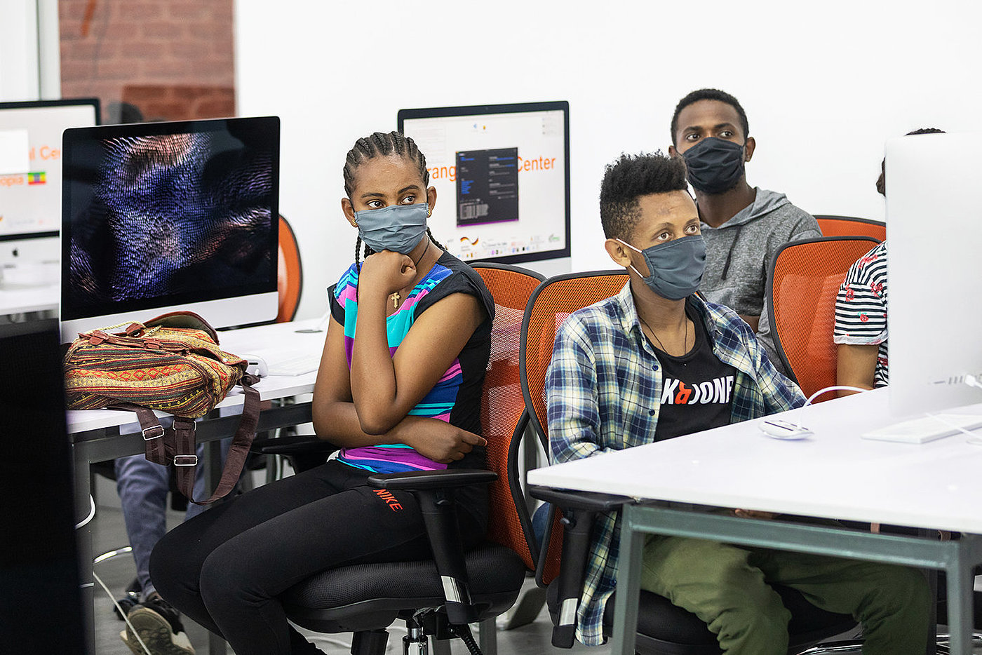 Photo: Several people wearing surgical masks sit at computer workplaces. All of them are looking in the same direction.