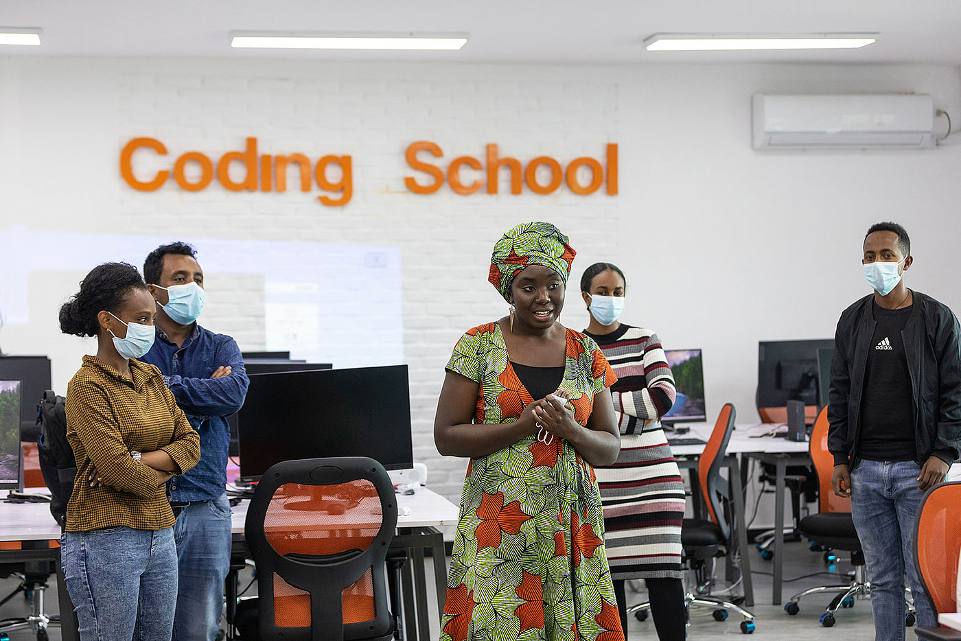 Photo: A woman is standing in the middle of a room and speaking. Other people stand around her in a circle; they are all wearing surgical masks. In the background we can read ‘Coding School’ on the wall.