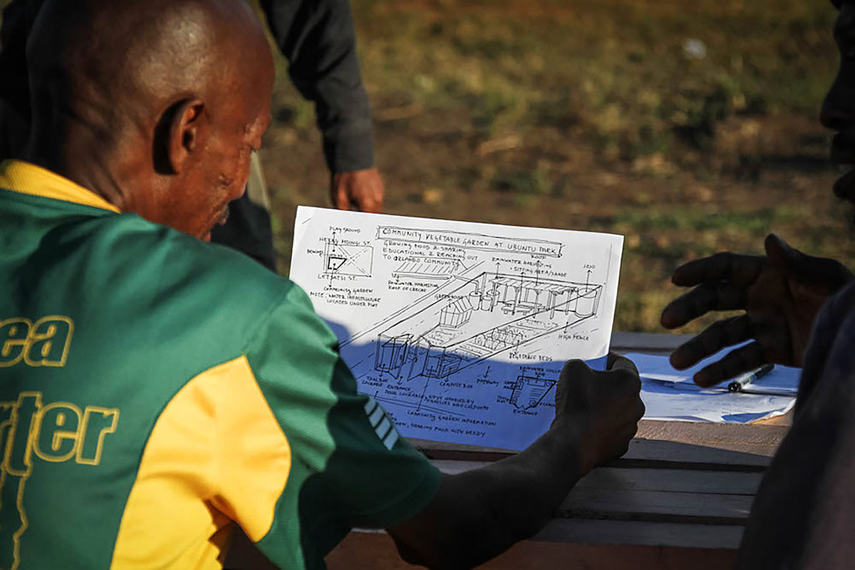 Photo: Several people stand or sit around a wooden table outside. One person is holding sketched technical plans in both hands and looking at them.