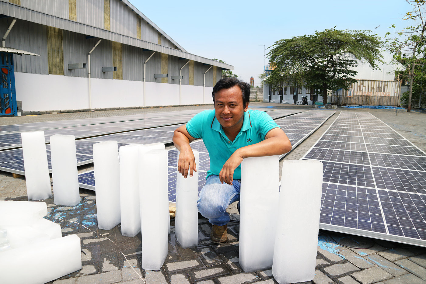 Photo: A courtyard with a large number of solar panels lying flat on the ground. A man is crouching next to them, leaning on small blocks of ice.