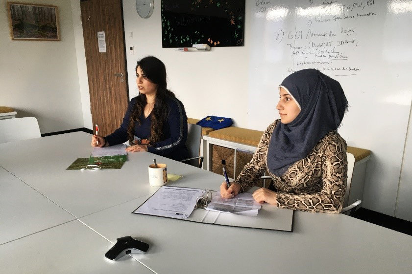 Photo: Two women sit at tables in a training room. Both are taking notes.