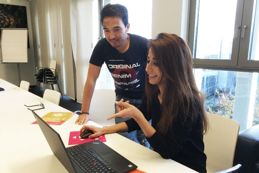 Photo: Two people at a table with an open laptop. One person is using the laptop, while another person stands beside the table.