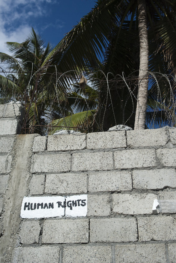 Photo: A wall topped with barbed wire, and high palm trees in the background. The words ‘Human rights’ are written on the wall.