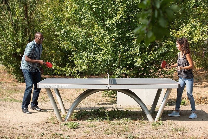 Photo: Two people play table tennis with trees in the background.