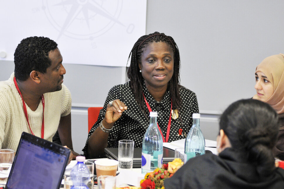 Photo: Four persons are sitting at a table with laptops, documents and drinks. One woman is talking and the others are looking at her.