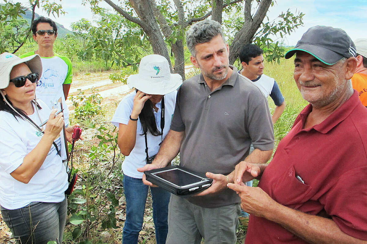 Photo: Several people standing in a field in front of a tree. A man is holding a tablet in his hands.