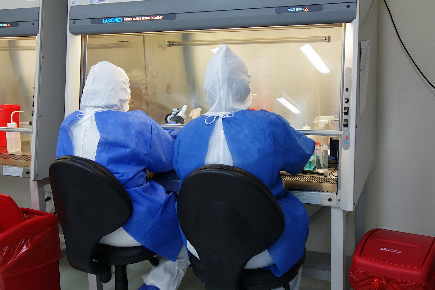 Photo: Two people in protective clothing working with their hands in a small laboratory with a glass front.