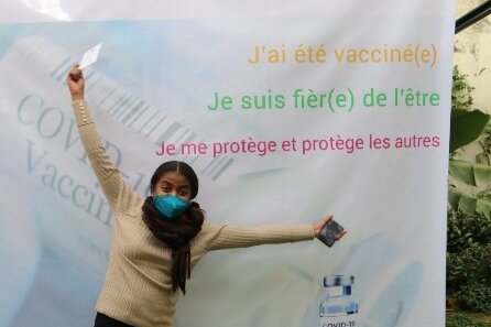 Foto: Eine junge Frau mit Mundschutz streckt die Arme hoch. Sie steht vor einem Plakat mit der Aufschrift: J’ai été vacciné(e). Je suis fièr(e) de l’être. Je me protège et protége les autres.
