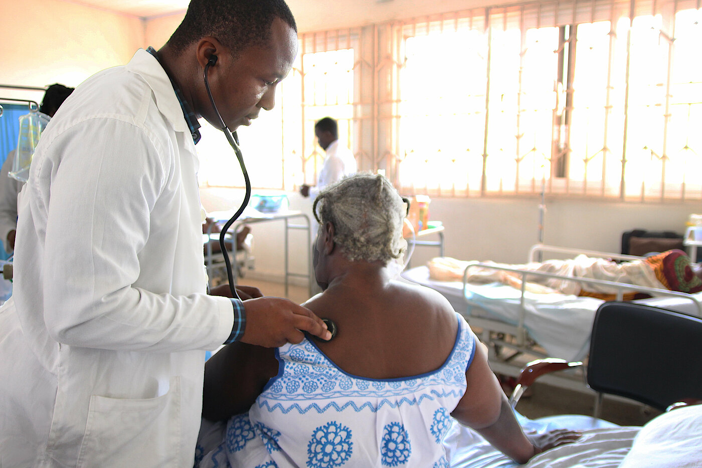 Photo: A doctor listening to a women’s back through a stethoscope in a doctor’s room. There are other people in the background.