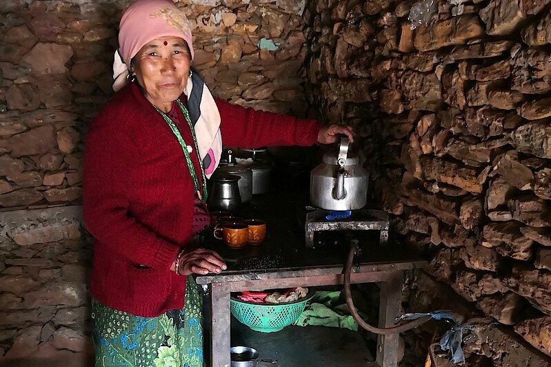 Photo: A woman standing in front of a stone wall at a cooking stove holding a pot of water on a gas hob.