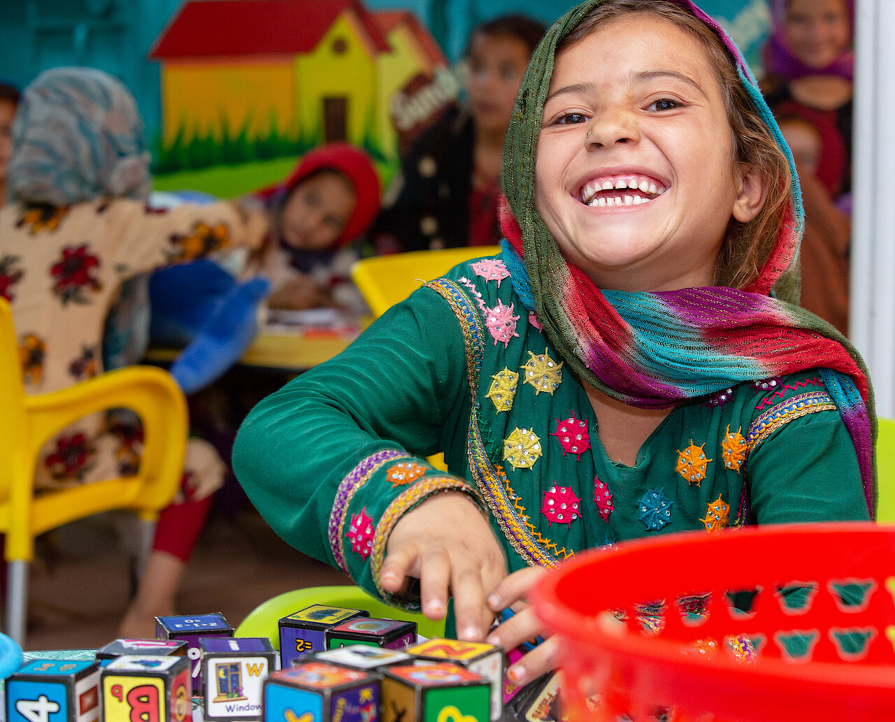 Photo: A girl with a colourful dress and scarf smiling at the camera.