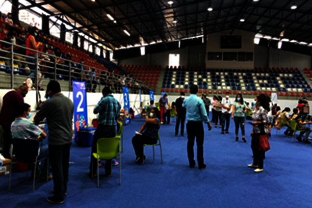 Foto: In einer großen Halle sitzen Menschen auf Stühlen, einige stehen in einer Schlange. Alle tragen Mundschutz.