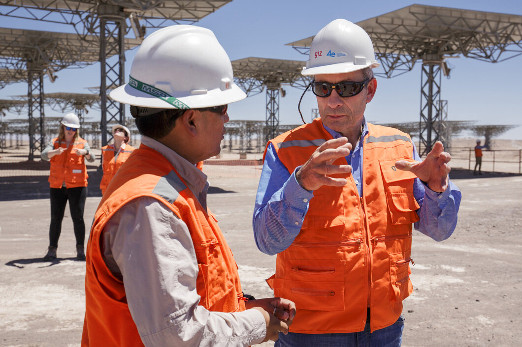 Two men wearing high-visibility vests and helmets talking to each other. There are more people in high-visibility vests and helmets in the background. They are all standing underneath a facility with a number of platforms on top of lattice towers.
