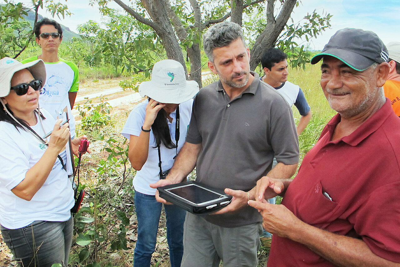 Photo: Several people standing in a field in front of a tree. A man is holding a tablet in his hands.