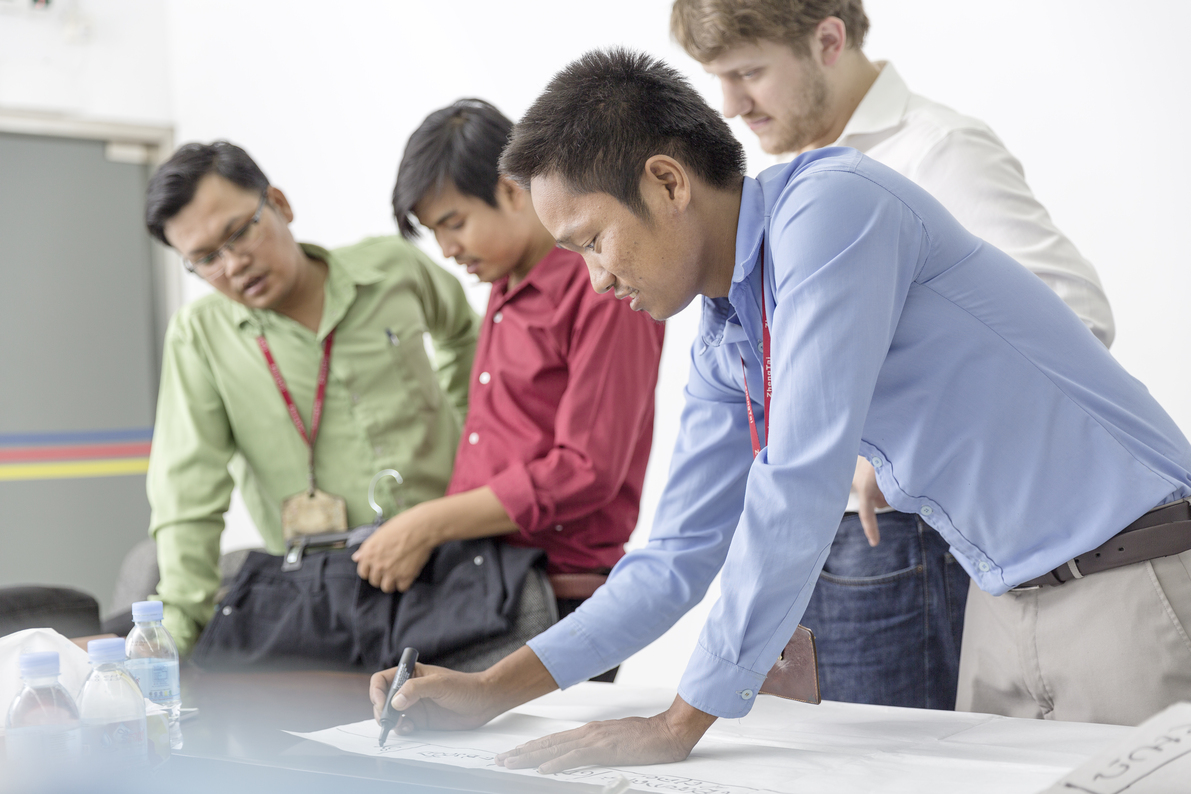 Photo: Four men standing at a table. One of them is writing something on a large piece of paper with a thick pen.