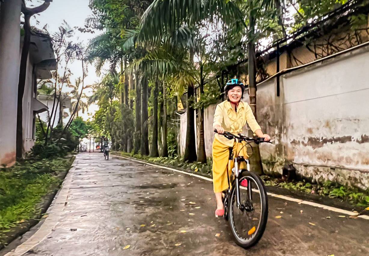 Photo: A woman wearing a helmet sits on a bicycle, with palm trees on the side of the road in the background.