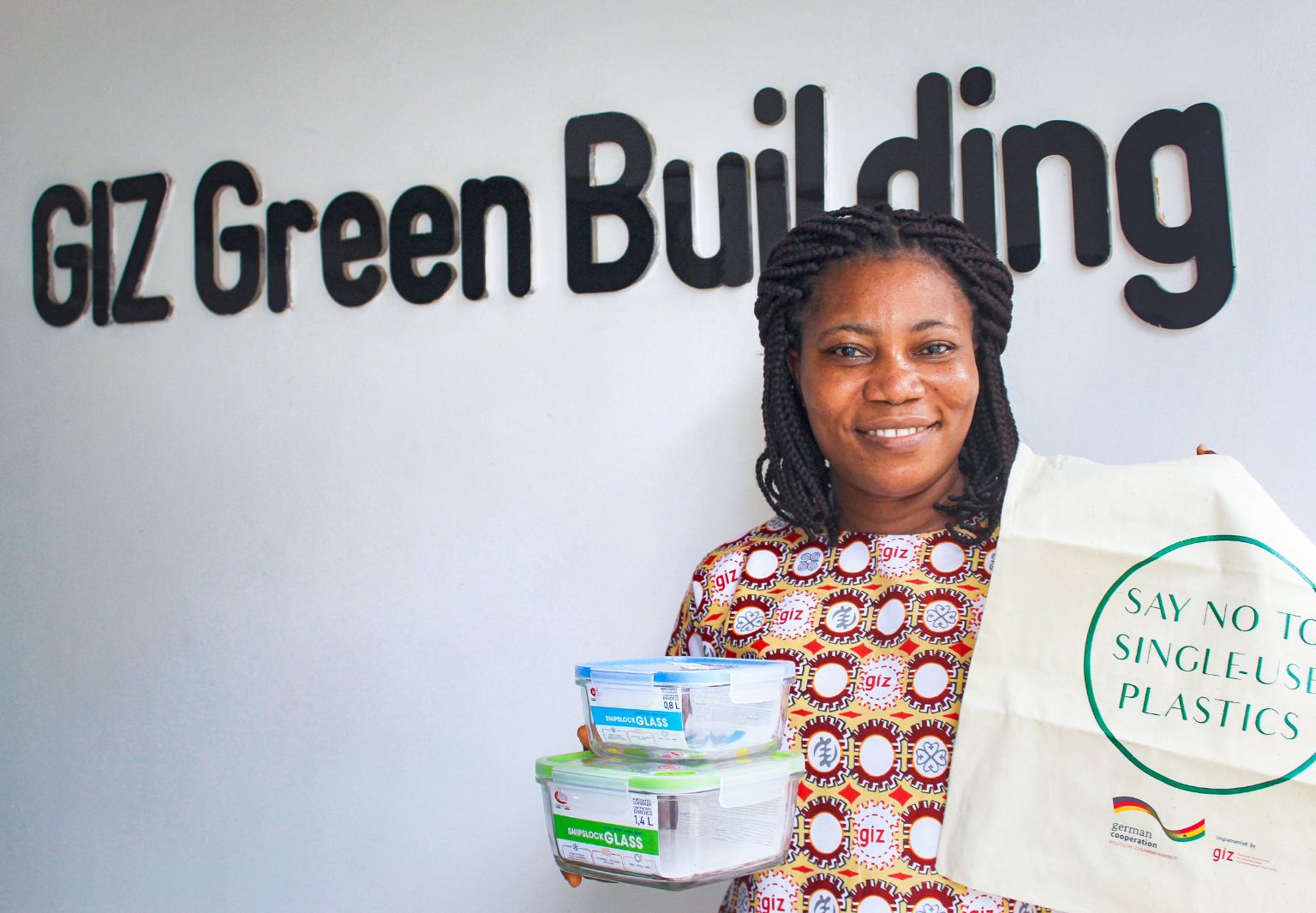 Photo: A woman standing in front of a wall with a sign that reads: ‘GIZ Green Building’. She is holding two glass containers with lids and a fabric bag printed with the words: ‘Say no to single-use plastics’.