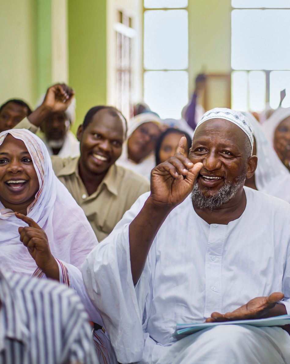 Photo: Several people are sitting in rows behind each other, some have raised their finger or hand. All are smiling.