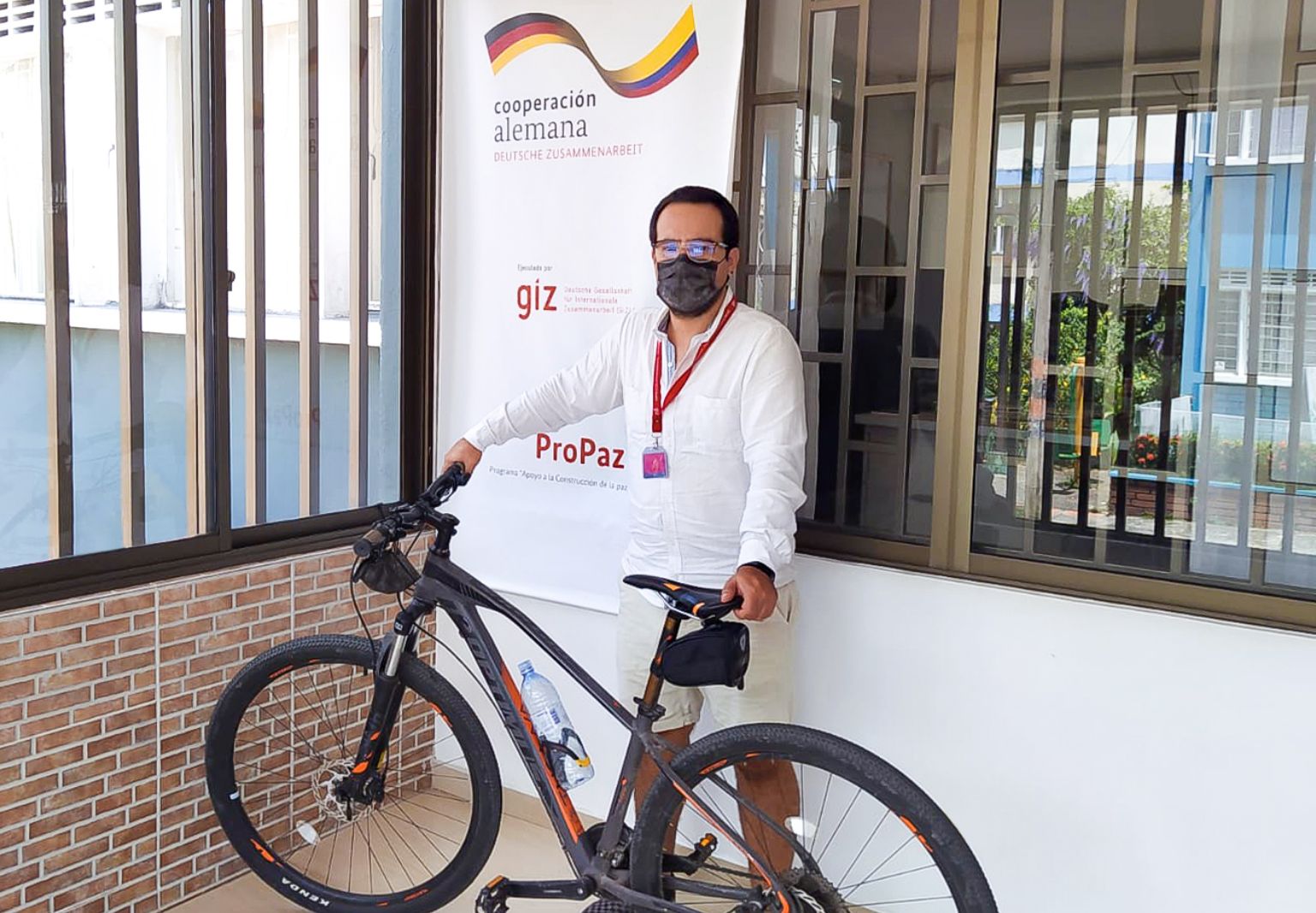 Photo: A man wearing a face mask standing in front of a building with the sign: ‘cooperación alemana, Deutsche Zusammenarbeit’ and the GIZ logo. A bicycle stands in front of him, which he is holding with both hands.