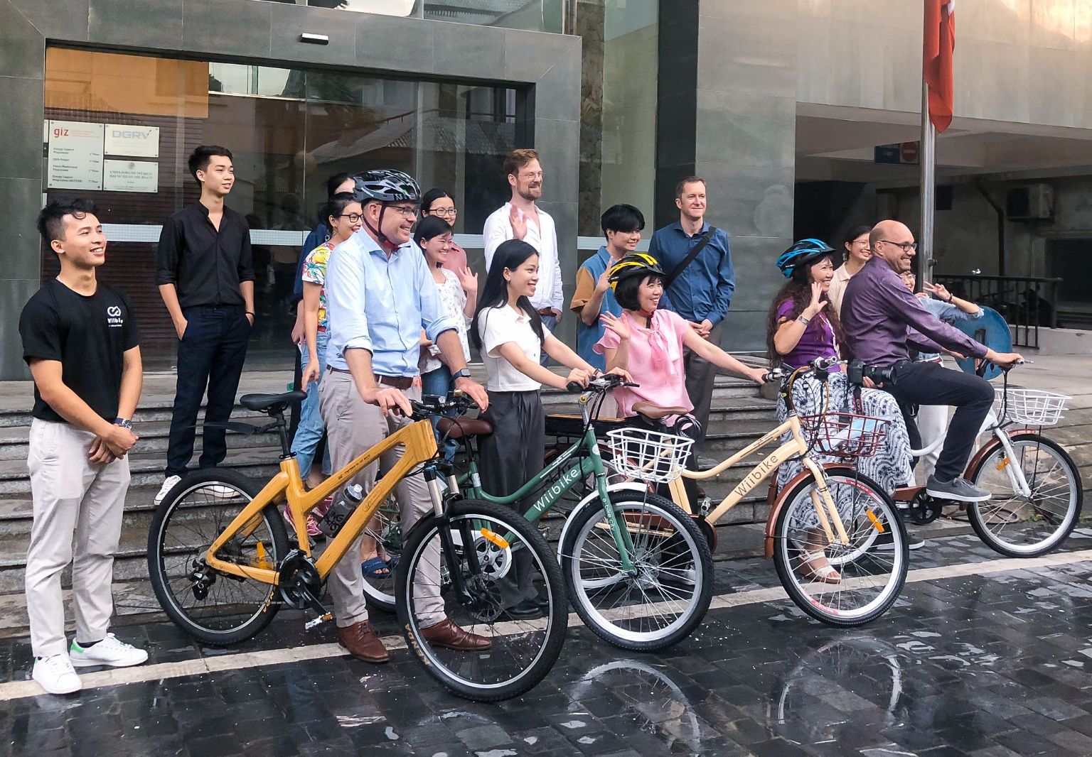 Photo: A group of people, some wearing bicycle helmets, standing in front of a building with the GIZ logo. Everyone is smiling, some are waving. Four people are holding on to bicycles.