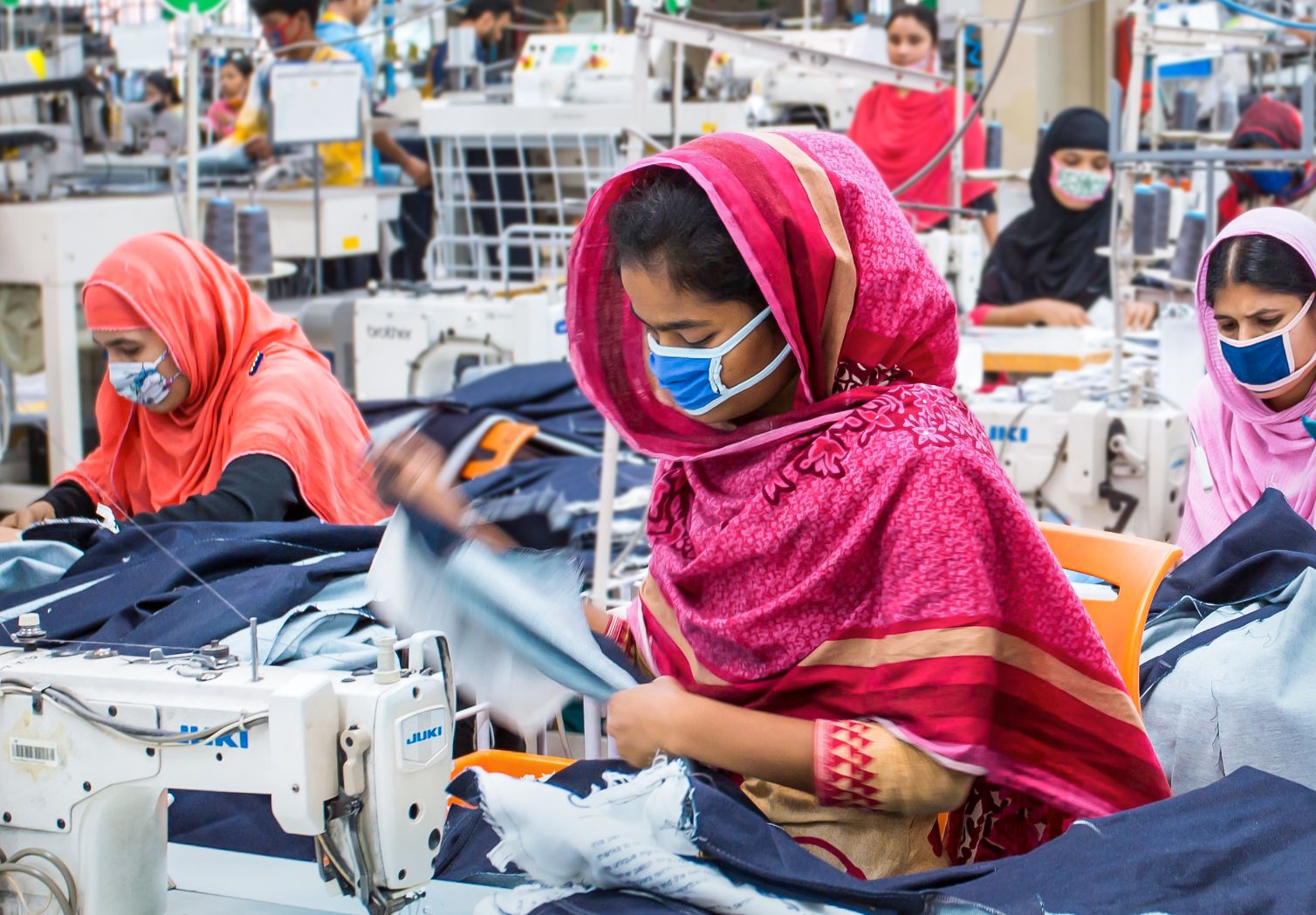 Photo: Seamstresses wearing headscarves and face masks sit in a large hall.