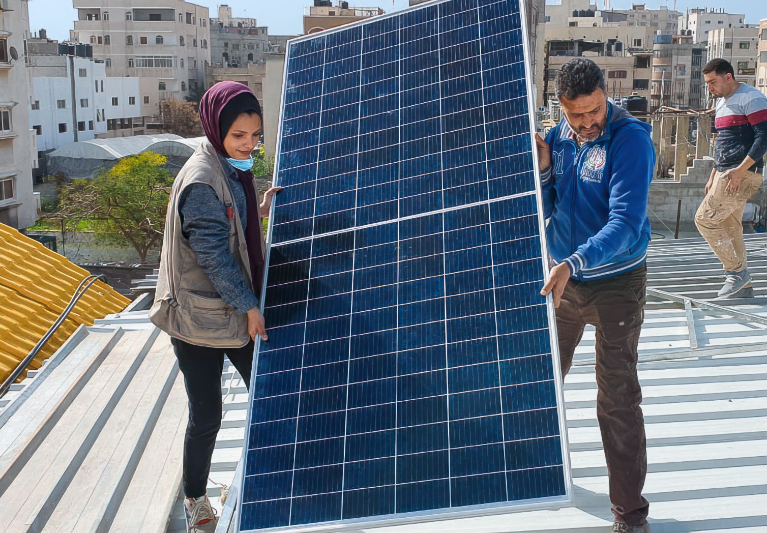 A woman with a headscarf and a man are on the roof of a house, carrying a large solar panel together.
