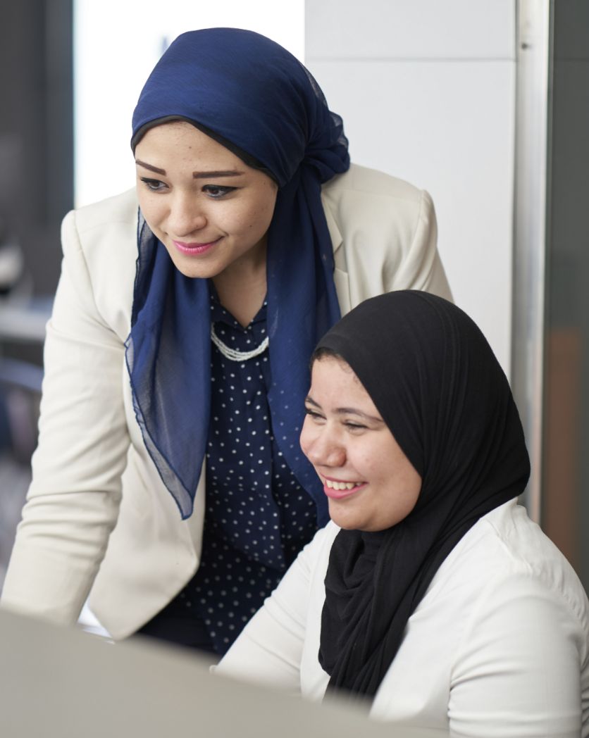 Photo: Two smiling women wearing headscarves and looking at a screen.