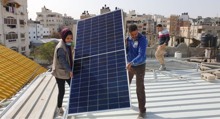 Photo: A man and a woman carrying a large solar panel across a roof.