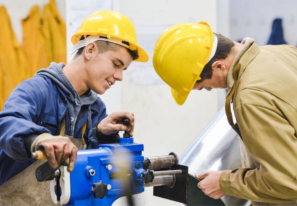 Photo: Two men wearing hard hats and working at a machine.