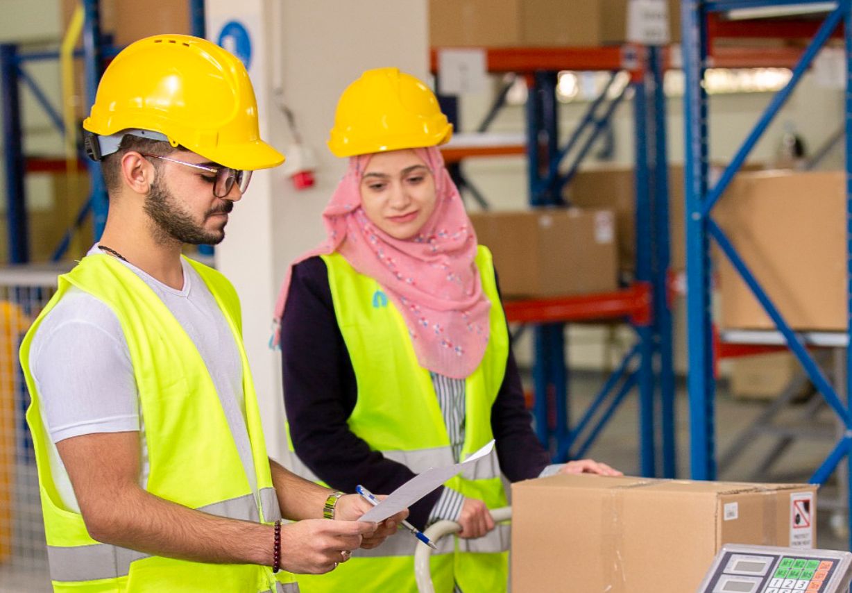 Photo: A woman and a man wearing hi-vis jackets and hard hats stand in a warehouse.