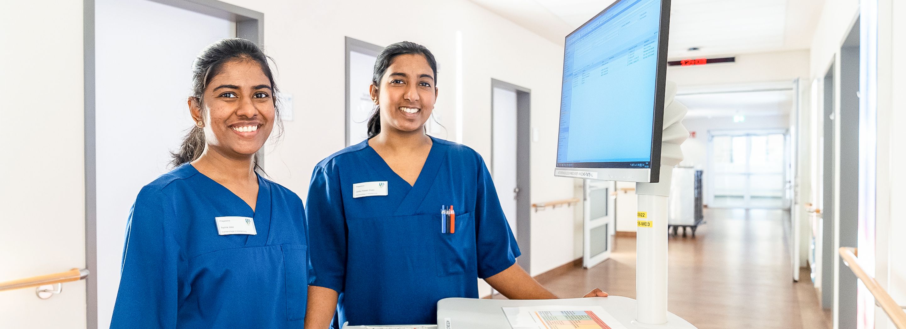 Photo: Two young women in dark blue scrubs standing in a hospital corridor next to a monitor.