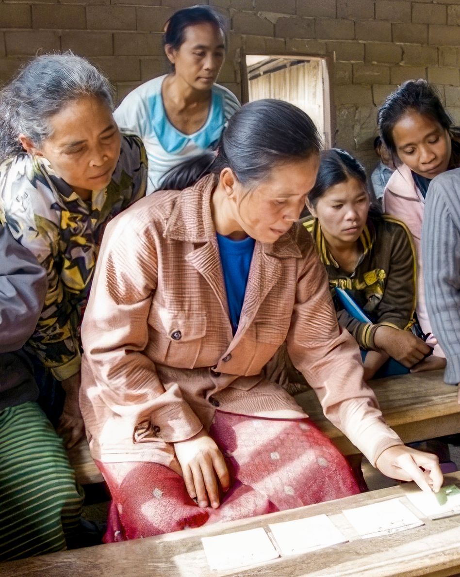 Photo: A group of women gathers around a wooden workbench with sheets of paper on it. The woman at the front is gesturing at one of the sheets.